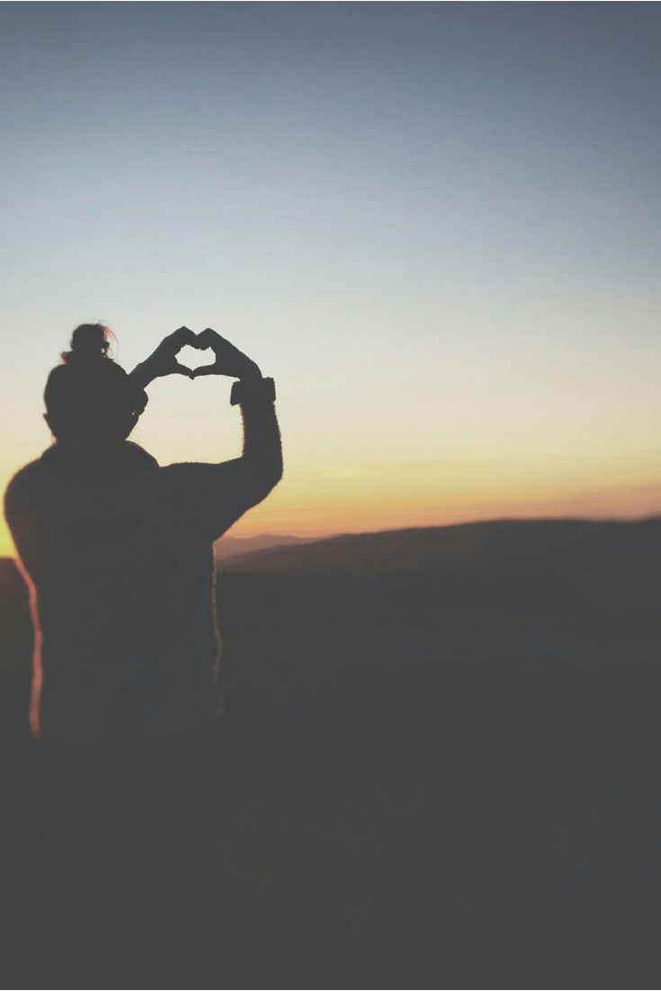 una mujer frente a una puesta de sol haciendo un corazón con las manos